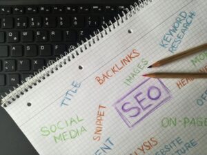 Notebook page with colorful handwritten SEO terms like backlinks, keyword research, social media, and images, placed on a laptop keyboard with pencils.