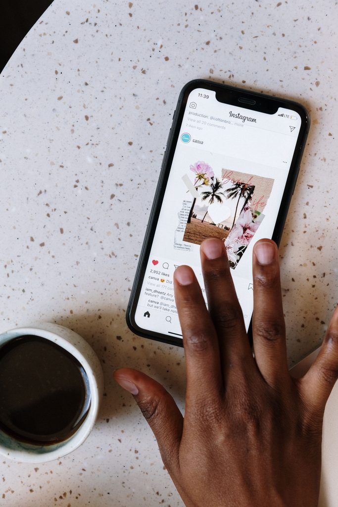 Hand tapping an Instagram post from Canva on a smartphone next to a cup of coffee.